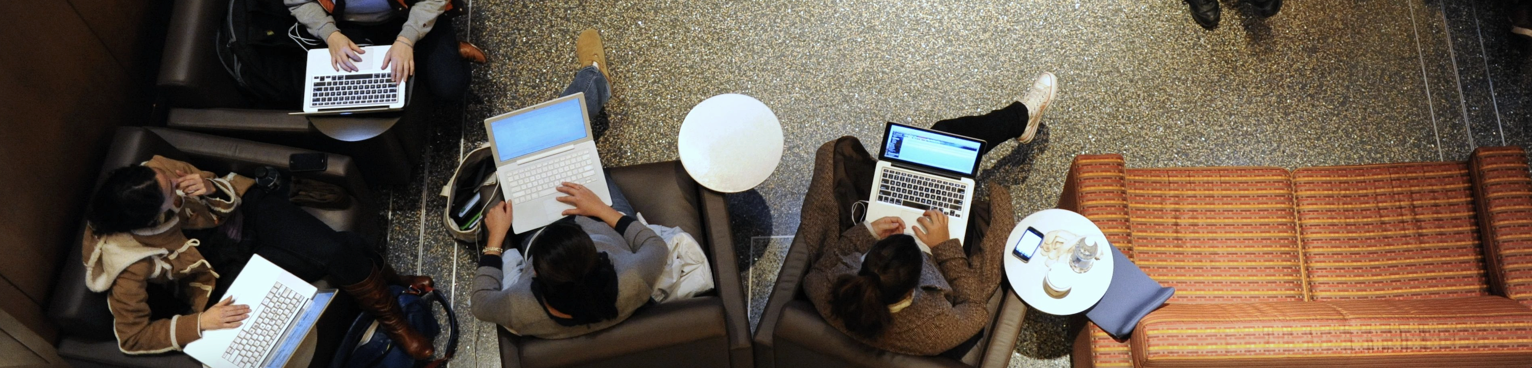 A small group of people sitting in a lounge area using laptops, viewed from above. There are armchairs, a sofa, and small round tables.